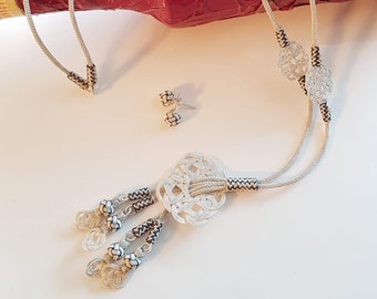 May include: A silver necklace with a large, intricate knot pendant and three smaller knot charms. The necklace is made of two strands of cord with black accents. The necklace is displayed on a red background.