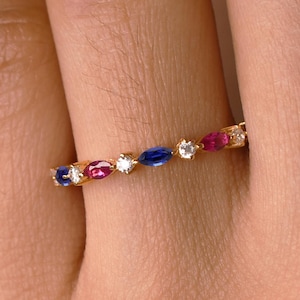 May include: A gold band ring with alternating red, blue, and white gemstones. The ring features oval-cut gemstones, creating a colourful and elegant design. The ring is worn on a finger.