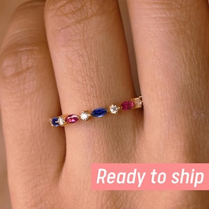 May include: A gold ring with alternating red, blue, and white gemstones. The ring is set with marquise-cut stones, creating a colorful and elegant design. The ring is shown on a finger.