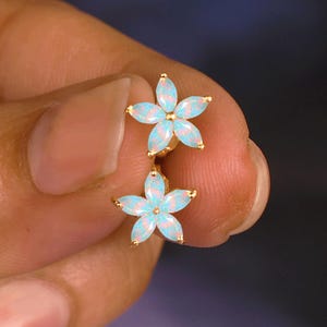 Puede incluir: Un par de pendientes de botón con forma de flor dorada. Cada pendiente presenta una piedra de ópalo azul claro y blanco dispuesta en un diseño de flor de cinco pétalos. Los pendientes se sostienen en una mano sobre un fondo oscuro.