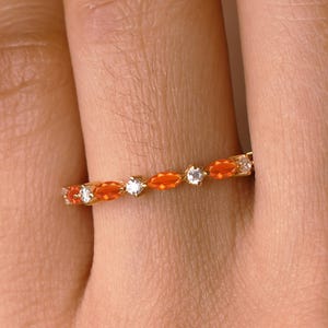 May include: A gold ring featuring alternating orange and clear gemstones. The gemstones are set in a row around the band. The ring is shown on a person's finger.