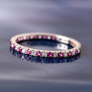 Puede incluir: Un delicado anillo de eternidad con una fila continua de piedras preciosas rosas y blancas alternadas. El anillo está hecho de metal blanco y tiene un acabado pulido.