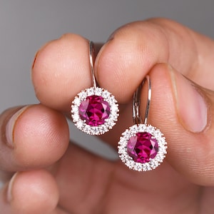 May include: A pair of silver earrings with a round, red gemstone surrounded by smaller, clear gemstones.