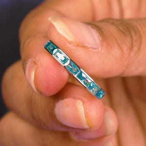 以下が含まれることがあります： 長方形の、薄い青色の宝石が並んだ銀色の指輪。指の間で指輪が持たれており、宝石の色と指輪のデザインが際立っています。この指輪はジュエリーです。