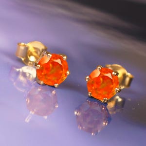 Może przedstawiać: Para złotych kolczyków sztyftów z błyszczącymi pomarańczowymi kamieniami szlachetnymi. Kamienie są oszlifowane, aby odbijać światło. Kolczyki leżą na odblaskowej, fioletowej powierzchni.