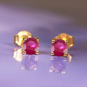 May include: A pair of gold stud earrings, each featuring a vibrant, round, ruby-red gemstone. The earrings are set against a soft, blurred purple background, highlighting the jewelry's sparkle and color.