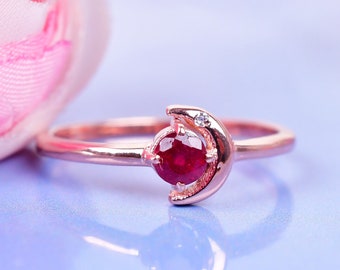 Könnte beinhalten: Ein Ring aus Roségold mit einem Halbmond-Design und einem roten Rubin-Edelstein. Der Ring liegt auf einem hellblauen Hintergrund.