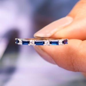 May include: A silver ring featuring alternating rectangular blue and small white gemstones. The ring is held between fingers, highlighting the details of the jewellery. The ring's design is simple and elegant, suitable for everyday wear or special occasions.