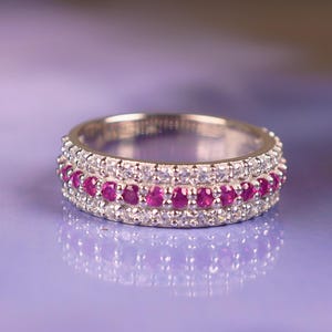 Pode incluir: Um anel de prata com três fileiras de pedras preciosas. As fileiras superior e inferior são incrustadas com pequenas pedras transparentes, enquanto a fileira do meio apresenta uma série de pedras preciosas vermelhas vibrantes. O anel está sobre um fundo roxo suave.