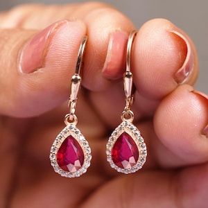 Op de afbeelding: Een paar roségouden oorhangers. Elke oorbel heeft een druppelvormige, dieprode edelsteen, omringd door een halo van kleine, heldere stenen. De oorbellen hebben een klapsluiting.