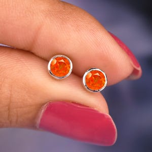 May include: A pair of round stud earrings featuring vibrant orange fire opal gemstones set in polished silver-toned metal bezels. Each circular stone displays a faceted surface that catches the light, creating a bright and reflective appearance.