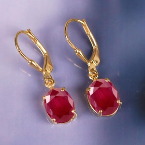 May include: A pair of gold-colored leverback earrings, each featuring a large, oval-cut, deep red gemstone. The gemstones are set in a gold-colored bezel setting. The earrings are displayed against a blurred, blue and purple background.