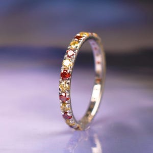 May include: A silver ring featuring a band of alternating yellow, red, and orange gemstones. The ring is set against a blurred background of purple and blue hues, with a reflective surface.