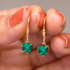 May include: A pair of gold-coloured leverback earrings, each with a heart-shaped, emerald-green gemstone. The earrings are shown against a neutral background.