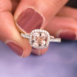 May include: A square-cut, peach-colored gemstone ring with a halo of smaller, clear stones. The ring's band is also adorned with small, clear stones. The ring is held up by a hand with painted nails.