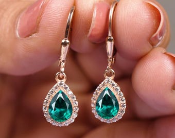 Puede incluir: Un par de pendientes colgantes dorados. Cada pendiente presenta una piedra preciosa verde esmeralda en forma de pera, rodeada por un halo de pequeñas piedras claras. Los pendientes se sujetan con un cierre de palanca.