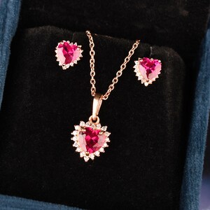 Puede incluir: Un conjunto de joyería compuesto por un collar y dos pendientes, cada uno con una piedra preciosa de color rosa oscuro en forma de corazón rodeada por un halo de pequeñas piedras claras, engastado en metal tono oro rosa con cadena.
