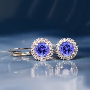 Puede incluir: Un par de pendientes de plata con una piedra preciosa redonda azul rodeada por un halo de piedras preciosas más pequeñas y transparentes.