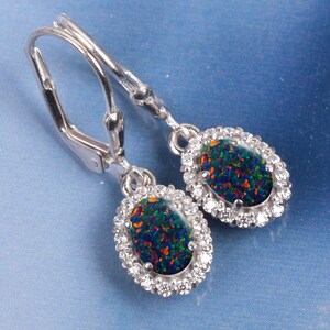 May include: A pair of silver leverback earrings featuring oval-shaped black opals with vibrant red and green flashes. Each opal is surrounded by a halo of small, clear, round-cut stones, creating a classic and elegant jewelry design.