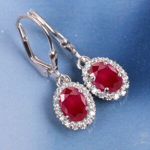 May include: A pair of silver leverback earrings featuring oval-cut deep red rubies as the center stones. Each ruby is surrounded by a halo of small, clear, round-cut white stones, creating a classic drop design with a polished metallic finish.