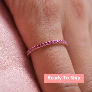 Puede incluir: Un delicado anillo de banda de oro con una fila de piedras preciosas rosas. El anillo está en un dedo y el texto "Ready To Ship" es visible en la imagen.