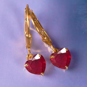 May include: A pair of gold-colored leverback earrings, each featuring a heart-shaped, faceted, deep red gemstone. The earrings are displayed against a soft, blurred background.
