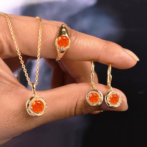 Op de afbeelding: Een goudkleurige sieradenset bestaande uit een ketting, een ring en een paar oorbellen met klapoorbelmechanisme. Elk sieraad heeft een ronde, feloranje edelsteen omringd door een halo van kleine, helderwitte steentjes in een gouden zetting.