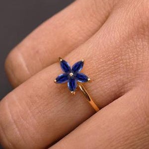 May include: A gold ring with a flower design, featuring five deep blue petals. The ring is set against a dark background, highlighting the contrast between the gold band and the blue gemstones. The ring is a piece of jewellery.