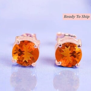 May include: A pair of stud earrings featuring round, faceted orange gemstones set in four-prong rose gold metal settings. The earrings are displayed with the text Jewel Bespoke visible near the right earring and Ready To Ship above.