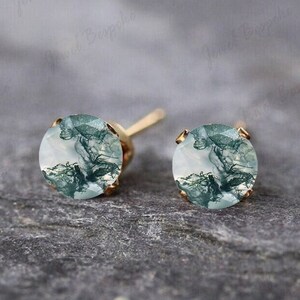 May include: A pair of round stud earrings featuring moss agate gemstones with green, fern-like inclusions set in gold-toned metal. Each circular stone displays unique, organic patterns of dark green mossy textures against a translucent white base.
