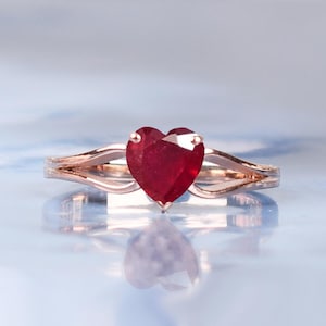 以下が含まれることがあります： ハート型の深紅色の宝石をあしらったローズゴールドの指輪。バンドは、中央の石を包み込む繊細な曲線を持つ、スプリットデザインです。柔らかく淡いブルーの背景に指輪がセットされています。