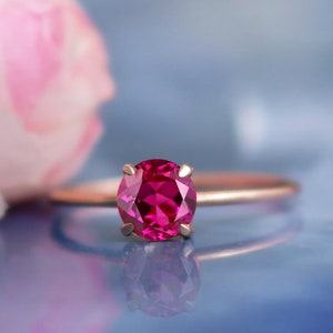 Może przedstawiać: Złoty pierścionek z różowym kamieniem szlachetnym. Pierścionek leży na niebieskim tle.