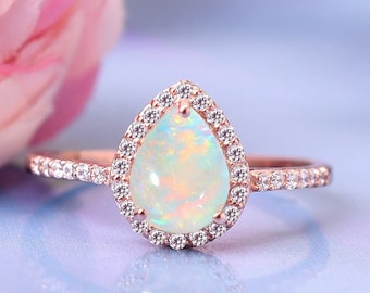 May include: A pear-shaped opal ring with a halo of small diamonds set in rose gold. The ring is on a light blue surface.