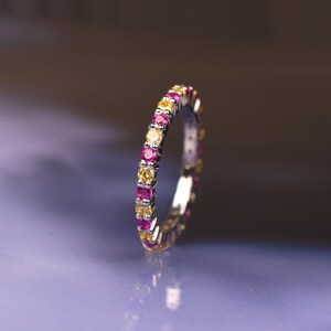 May include: A silver ring featuring alternating pink and yellow gemstones. The ring is adorned with small, round stones that encircle the entire band. The gemstones are evenly spaced, creating a continuous pattern of colour. The ring is displayed on a reflective surface.