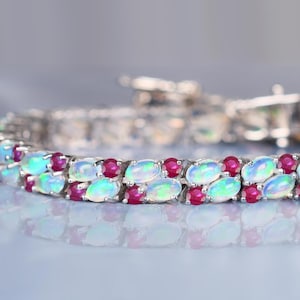 May include: A close-up of a silver bracelet featuring oval-cut opals and small, round rubies. The opals have a milky, iridescent quality, and the rubies provide a vibrant contrast. The bracelet is set on a reflective surface.
