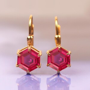 May include: A pair of gold earrings with hexagonal pink gemstones. The earrings have a simple, elegant design.