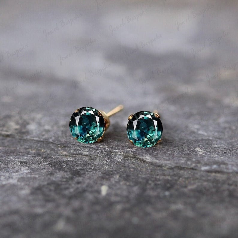 May include: A pair of gold stud earrings featuring round, teal-colored gemstones. The earrings have a classic design with a simple gold setting that complements the vibrant color of the stones. The gemstones have a faceted cut, enhancing their sparkle.