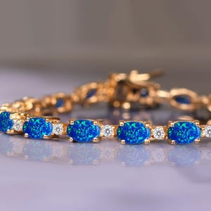 Puede incluir: Un brazalete dorado con piedras de ópalo azul ovaladas y pequeñas gemas transparentes. Los ópalos tienen un efecto brillante. El brazalete se muestra sobre una superficie reflectante.