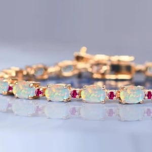 Puede incluir: Un brazalete dorado con ópalos ovalados y pequeñas gemas rojas. Los ópalos son de color blanco lechoso, y las piedras rojas añaden un toque de color. El brazalete se muestra sobre una superficie reflectante. Mide unos 18 cm.