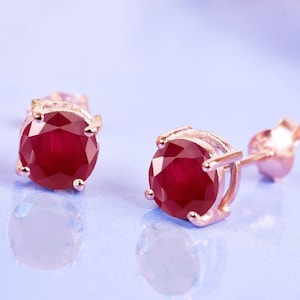 Pode incluir: Brincos de pressão com pedras preciosas redondas, vermelho escuro, cravejadas em ouro rosa. Os brincos são apresentados em uma superfície azul clara, destacando suas qualidades reflexivas e design elegante. As pedras são o foco.