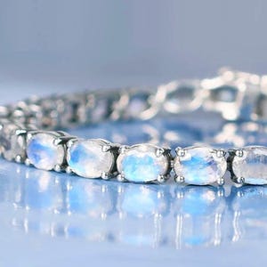 May include: A silver-toned tennis bracelet featuring a series of oval-cut moonstones. Each stone displays a translucent, milky white appearance with distinct blue adularescence. The stones are set in individual metal prongs along the chain.
