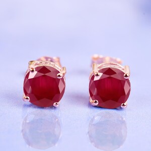 May include: A pair of round-cut red ruby stud earrings set in rose gold metal. Each earring features a faceted circular gemstone held in place by a four-prong setting, showcasing a deep crimson color and a polished metallic finish on the base.