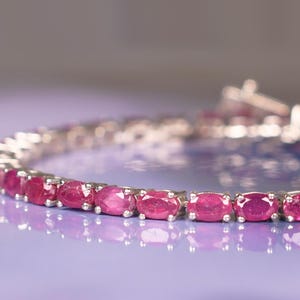 Pode incluir: Uma pulseira de prata com uma fileira de pedras preciosas rosa ovais. A pulseira está sobre uma superfície roxa.