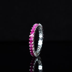 Può includere: Un anello a fascia d'argento con una fila di pietre preziose rosa a forma di baguette.