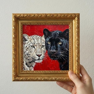 Puede incluir: Pintura enmarcada que presenta un leopardo y una pantera negra sobre un fondo rojo. El leopardo tiene pelaje blanco con manchas negras, mientras que la pantera es negra. El marco dorado tiene detalles ornamentales.