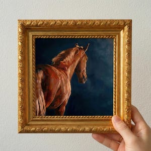 May include: Oil painting of a brown horse in a gold ornate frame. The horse is facing right, with its head turned towards the viewer. The background is a dark blue. The frame is held by a hand.