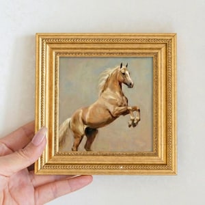 May include: A framed painting of a rearing palomino horse. The artwork features a golden frame and depicts the horse in shades of tan and white against a soft blue and grey background. The horse is posed with its front legs raised.