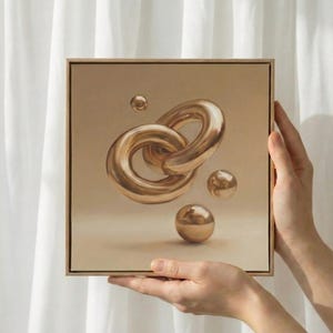 May include: A square canvas art piece featuring a gold-colored, linked chain design with three gold spheres. The artwork is held by two hands against a neutral background. The piece is framed in a light wood.