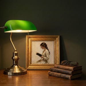 May include: A green-shaded desk lamp with a brass base and arm illuminates a framed painting of a woman reading. Beside the painting are a stack of old books and a pair of eyeglasses. The lamp is turned on.