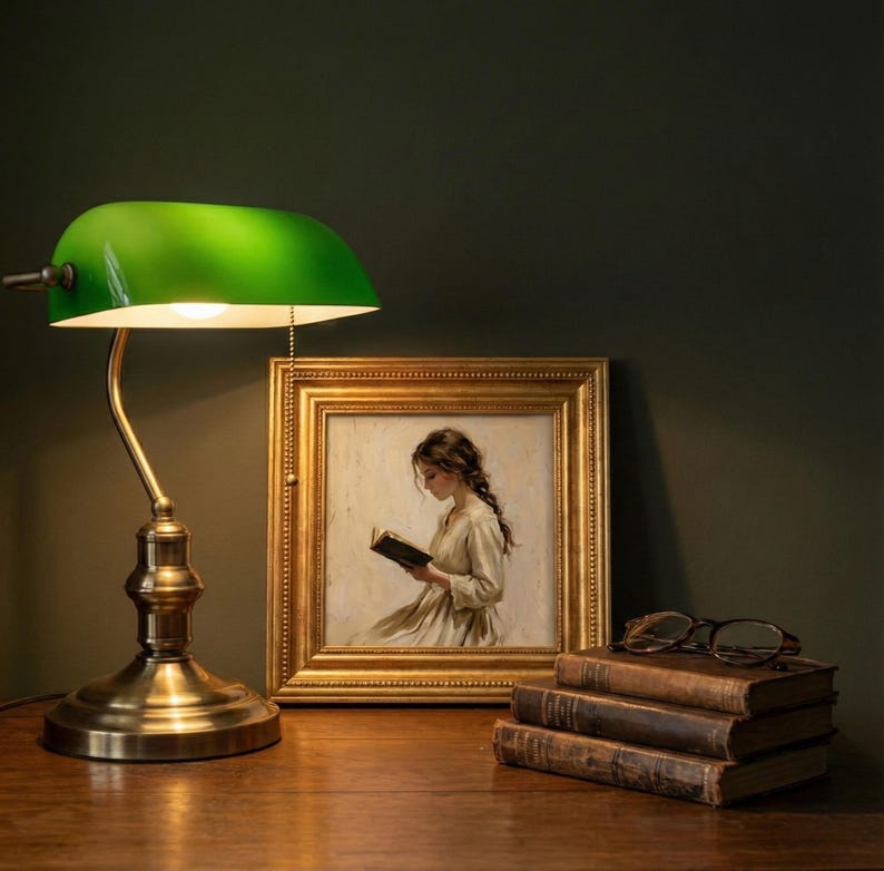 May include: A green-shaded desk lamp with a brass base and arm illuminates a framed painting of a woman reading. Beside the painting are a stack of old books and a pair of eyeglasses. The lamp is turned on.